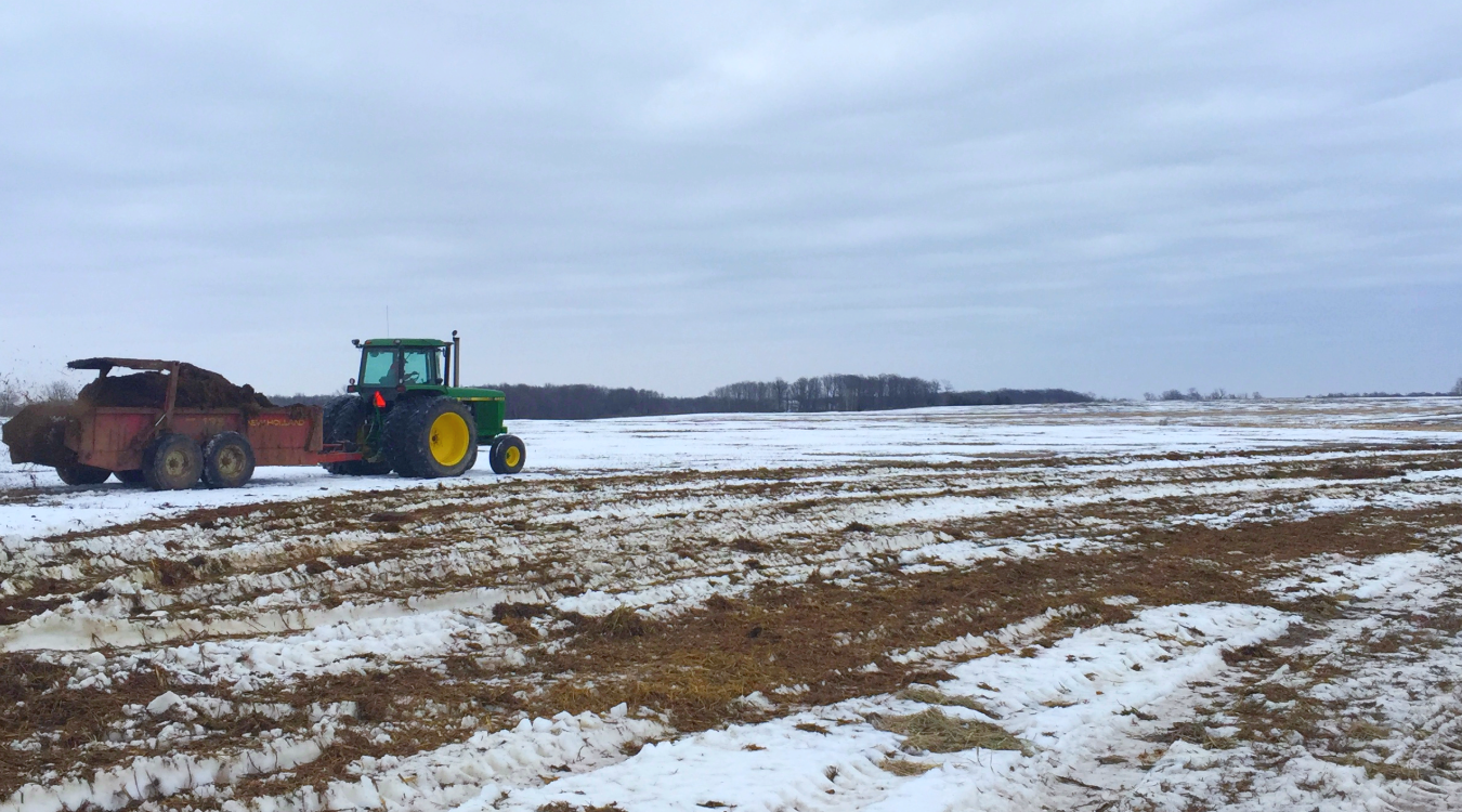 A tractor pulls a wagon spreading manure across a snow covered field