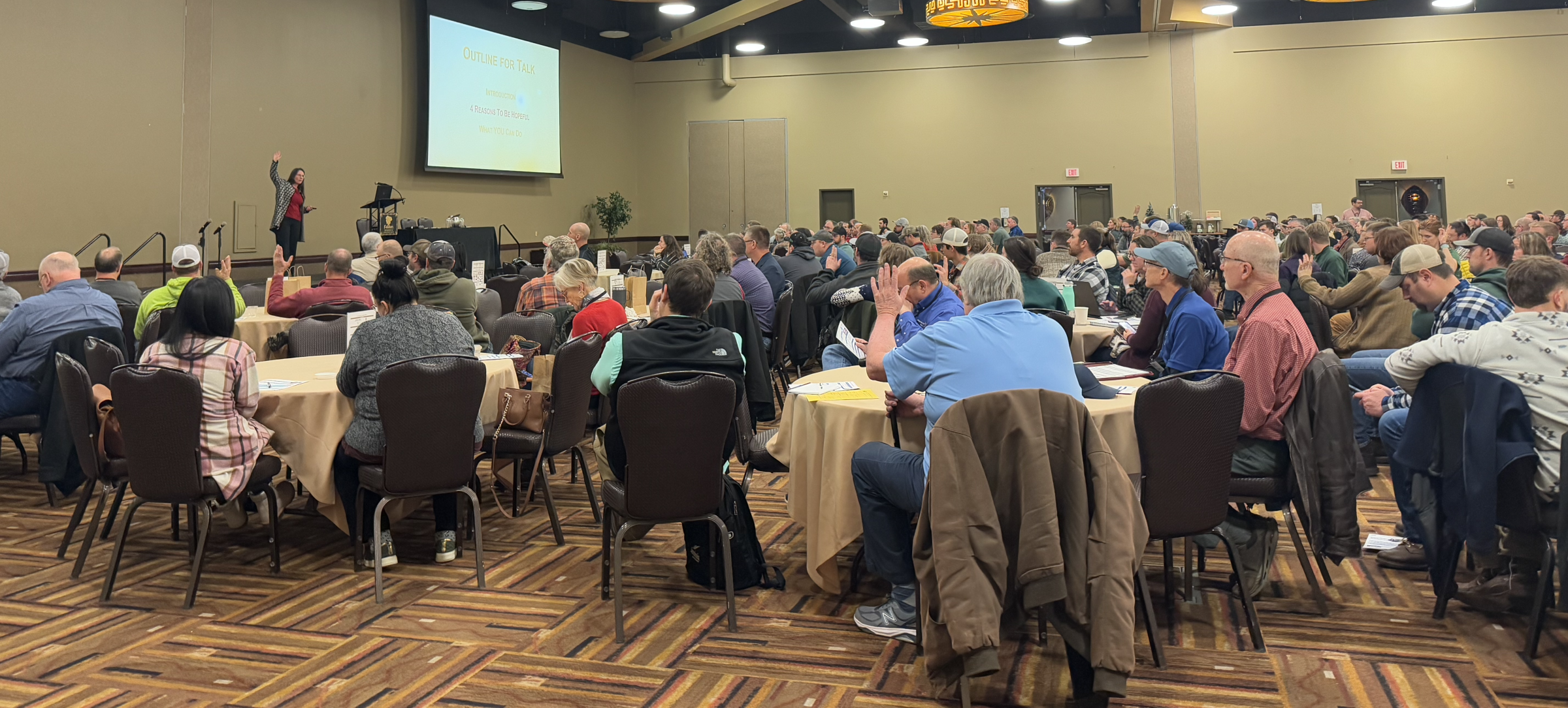 Large conference room with attendees at round tables, a presenter on stage, and a projection screen behind them.