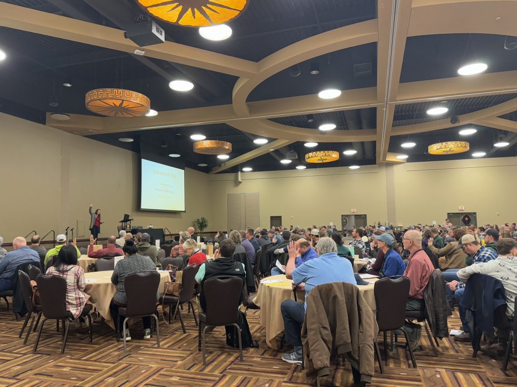 Large conference room with attendees at round tables, a presenter on stage, and a projection screen behind them.