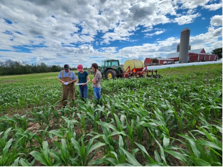 Three people examine corn field with farm equipment and red barn in background
