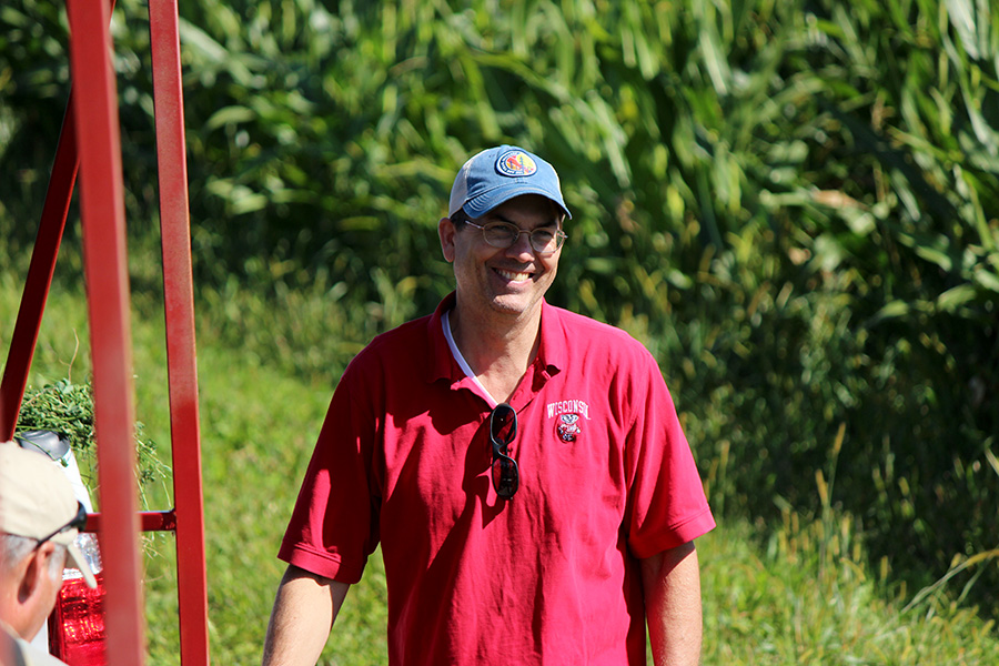 Man in red shirt and blue cap standing near tall green plants; another person partially visible on the left.