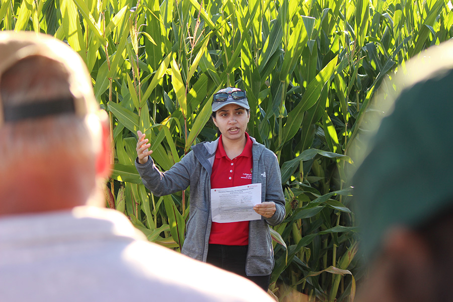 Woman speaking in front of a cornfield, holding paper, with two listeners in foreground.
