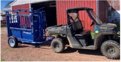 Utility vehicle towing a blue cattle chute near a red barn.