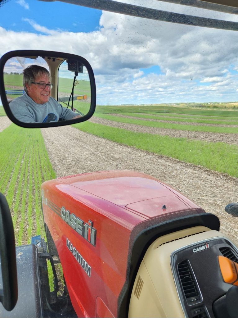 View from inside a tractor cab looking over the red hood at a striped field, with a Green Lake County farmer, Jim Hebbe, visible in the side mirror.