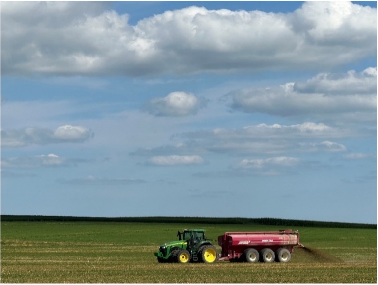 Green John Deere tractor pulling a red industrial agricultural tank spreader across a wide green and gold field under a cloudy blue sky.