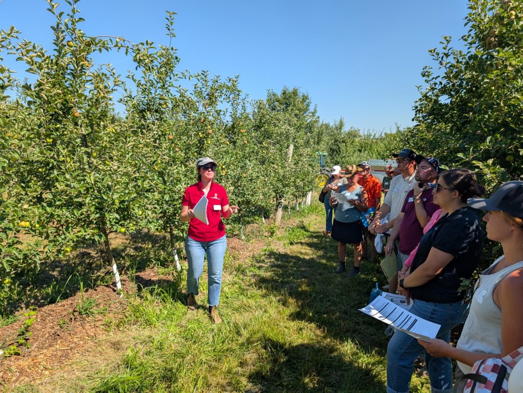 Amaya Atucha, in a red shirt, addresses a group of attendees standing among rows of apple trees in a sunny orchard.