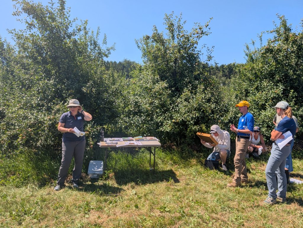 Hannah Gaines Day stands by a table with brochures in an apple orchard, speaking to a small group of attendees.