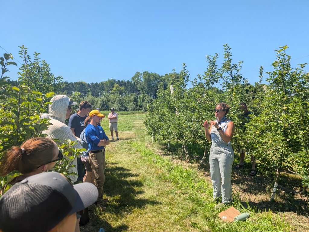 Leslie Holland, wearing striped overalls, speaks to a group of people standing between rows of apple trees.