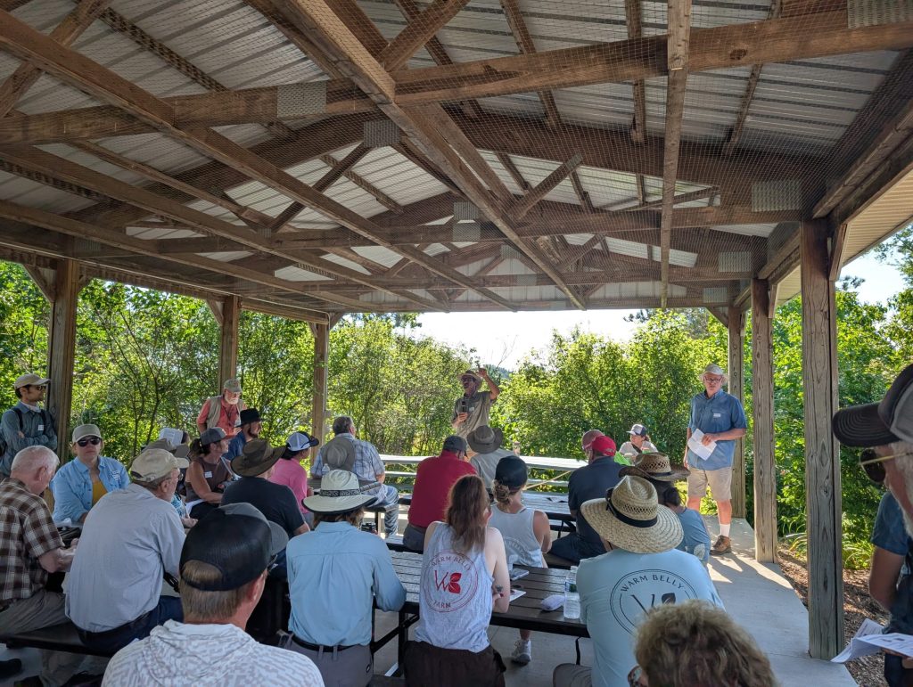 A group of people sits at picnic tables under a wooden open-sided pavilion, listening to presenters outdoors.