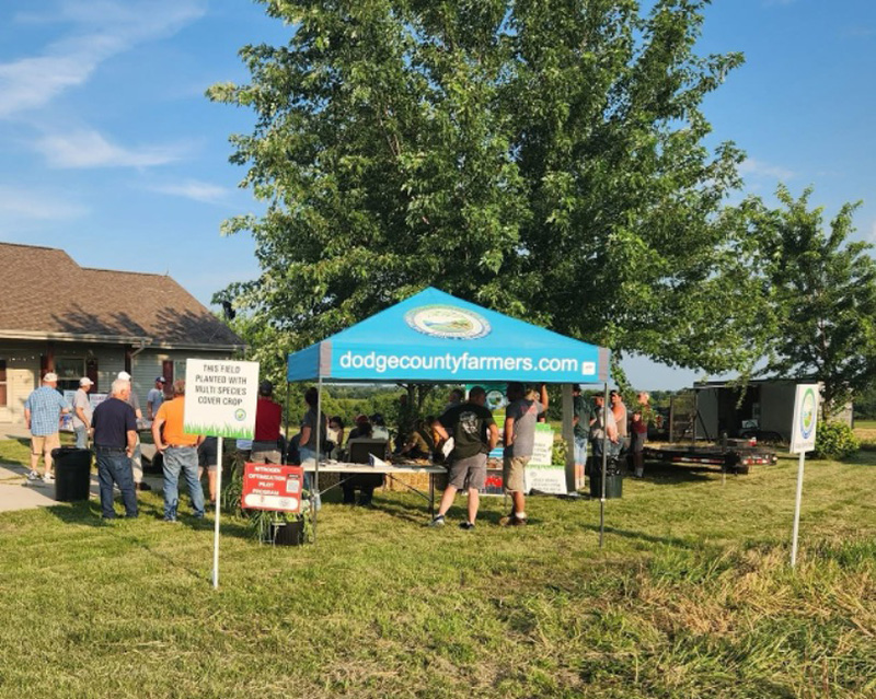 Attendees gather at an outdoor field day tent labeled “dodgecountyfarmers.com” beside crop demonstration signs.