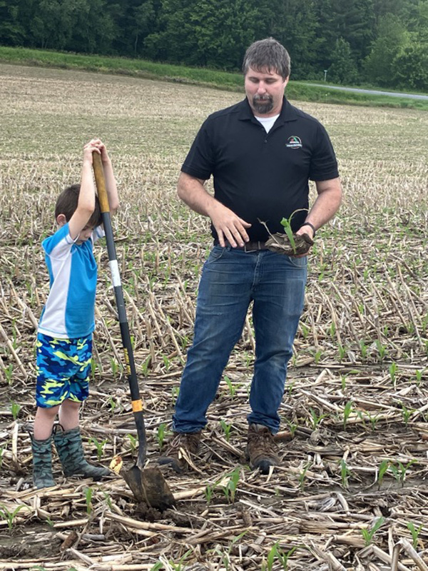 Person holds an uprooted corn plant while a child digs a soil pit with a shovel in a corn field.