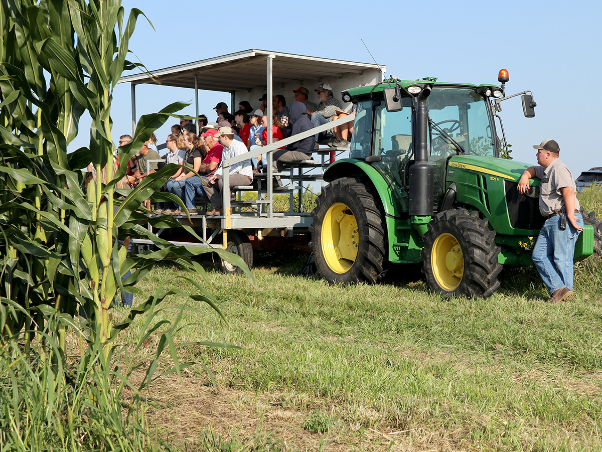 Field day attendees seated on a wagon beside a John Deere tractor, observing corn research plots in a field.